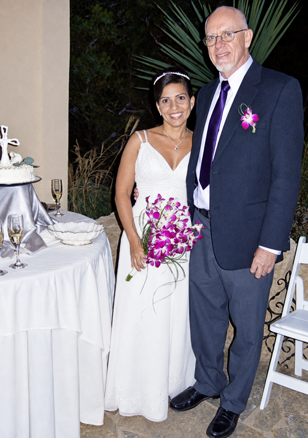 Pareja de recién casados junto a una mesa de celebración; la novia sostiene un ramo de orquídeas.